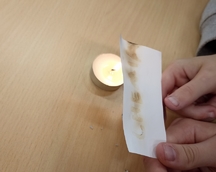 ,,Niewidzialne pismo" - na zdjęciu widać świeczkę i umieszczoną nad płomieniem kartkę, na której uwidacznia się napis ,,chemia". Wcześniej słowo to zostało zapisane za pomocą patyczka higienicznego i soku z cytryny.