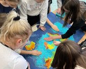 Dzieci wykonują konturową mapę świata  z wykorzystaniem barwionego ryżu. Na mapie zaznaczono wyzyny, niziny, góry  oraz morza i oceany.