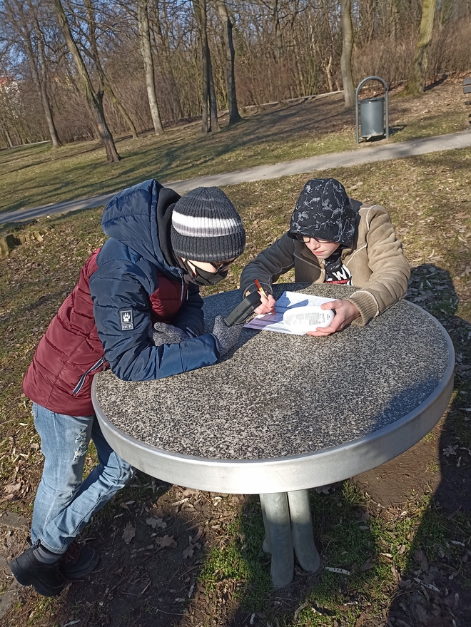 Dwóch chłopców w parku odczytuje dane z urządzeń pomiarowych. Jeden z nich stoi przy stoliku, drugi zapisuje wyniki.