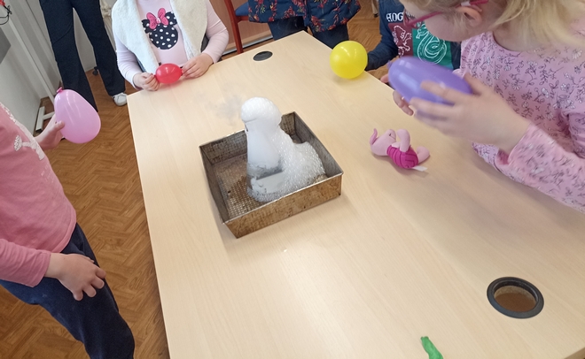 Na stole znajduje się tacka a w niej kolba stożkowa, do której wlano wrzątek i płyn do baniek, a następnie dodano kawałki suchego lodu. Młodsi uczestnicy trzymają baloniki, obserwują efekty przeprowadzonego eksperymentu.