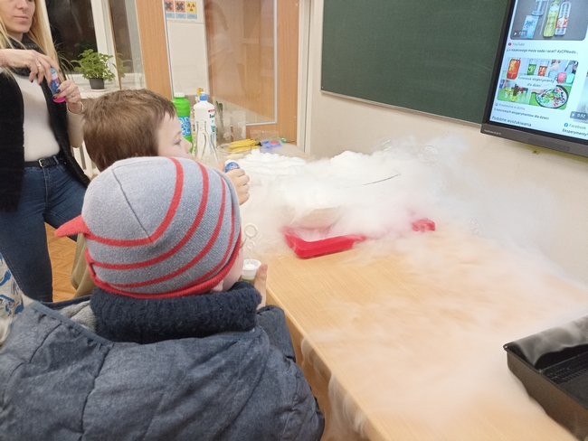Uczestnicy sprawdzają jak suchy lód reaguje na bańki mydlane. W tle tablica multimedialna, na której wyświetlone są zdjęcia ukazujące szkło laboratoryjne.
