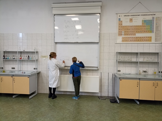 Uczeń zapisuje na tablicy reakcję chemiczną. Obok stoi pracownik Uniwersytetu Przyrodniczego, sprawdza poprawność zapisu.