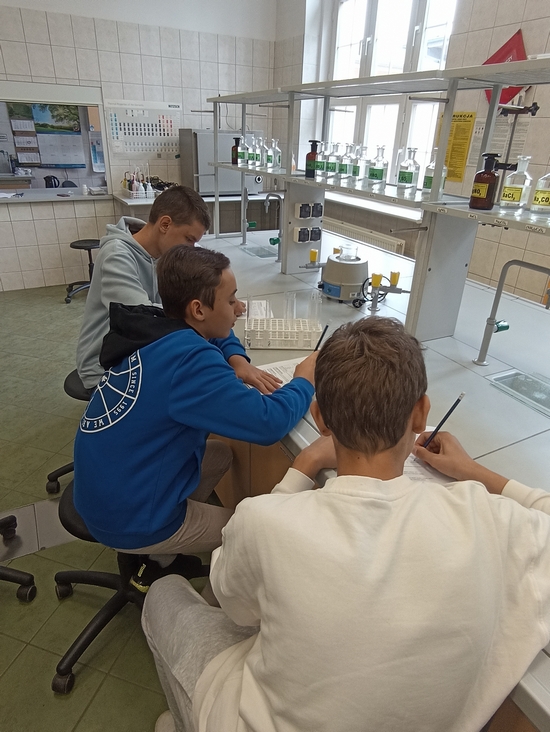 Uczniowie siedzą przy stołach laboratoryjnych i wykonują doświadczenia. Zapisują wyniki na kartach pracy.