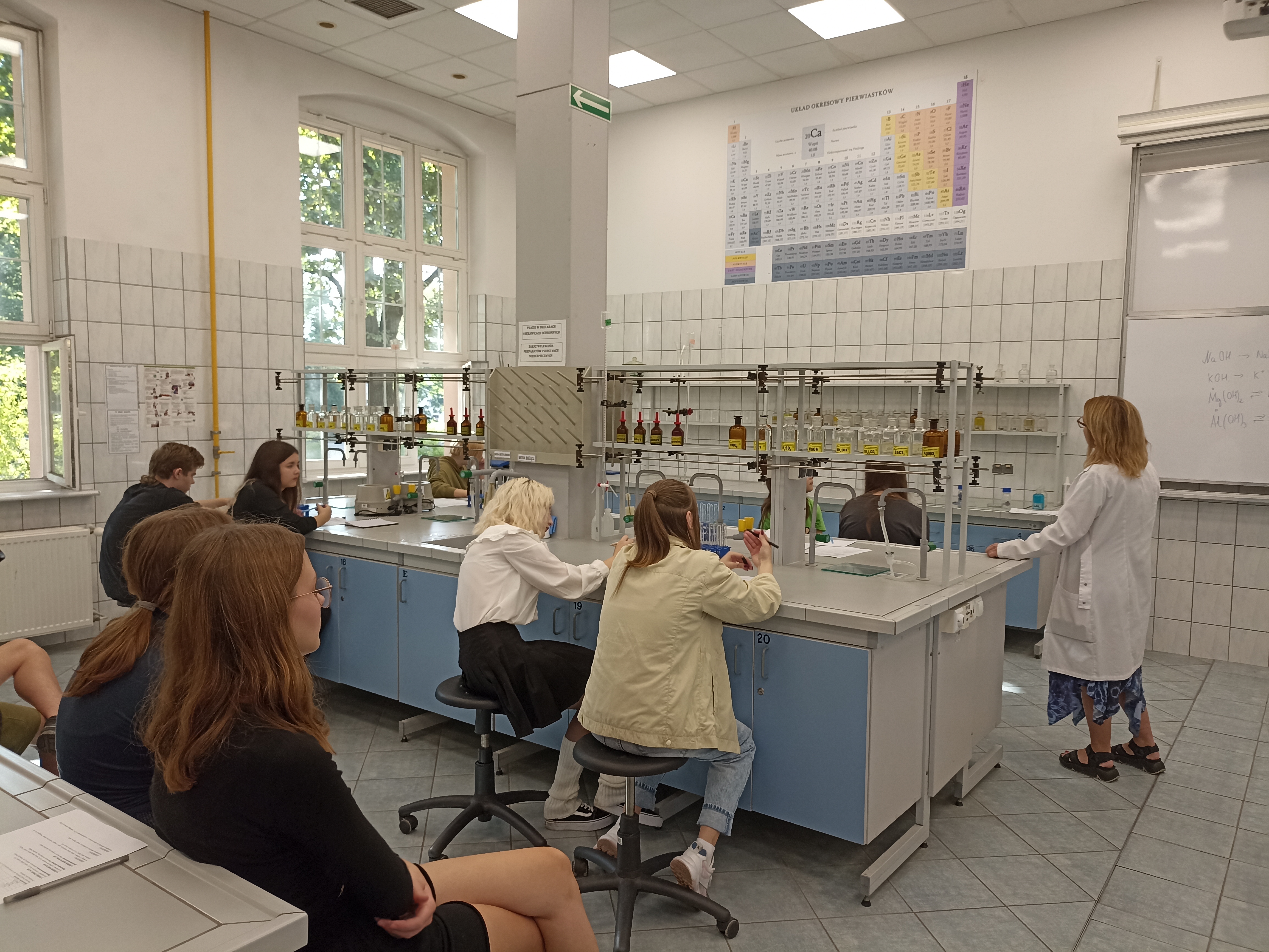 Uczniowie siedzą w grupach dwuosobowych przy stołach laboratoryjnych. Wszyscy wraz z nauczycielem Uniwersytetu Przyrodniczego wpatrują się w układ okresowy pierwiastków chemicznych.